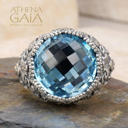 A sterling silver Byzantine filigree ring with a large blue topaz stone, set in a detailed filigree design on a rocky surface.