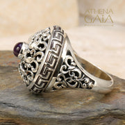 A silver ring featuring a Byzantine-inspired design with a purple amethyst stone, detailed with Greek keys and filigree work.