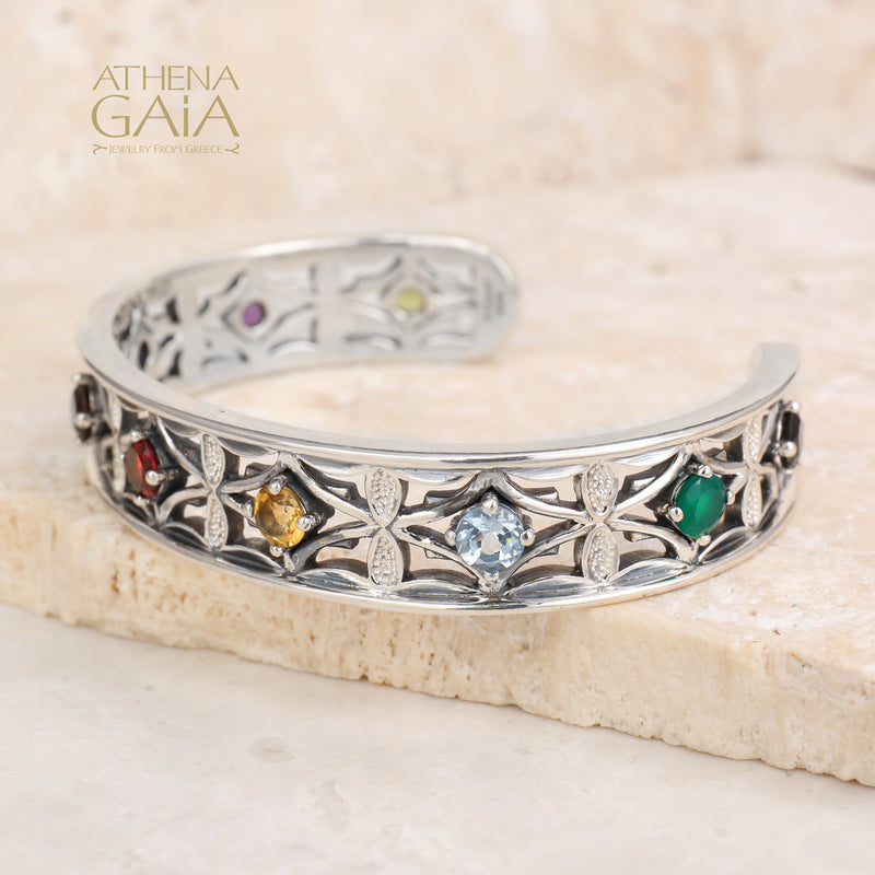 Silver ring with embedded gemstones on a beige stone surface, branded 'Athena Gaia'.