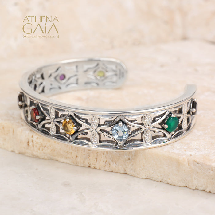Silver ring with embedded gemstones on a beige stone surface, branded 'Athena Gaia'.