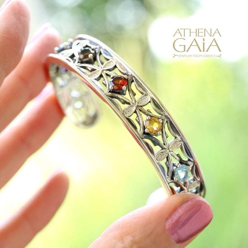 Silver bracelet with gemstones held by a hand against a blurred green background, featuring the Athena Gaia brand.