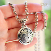 Hand holding silver pendant necklace with ornate cross pattern and bold chain in bright lighting.