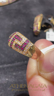 A woman's hand wearing a gold ring with a greek key geometric meander pattern of diamonds and rubies.