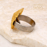 A gold-colored seashell ring displayed on a stone surface with the brand name Athena Gaia visible.