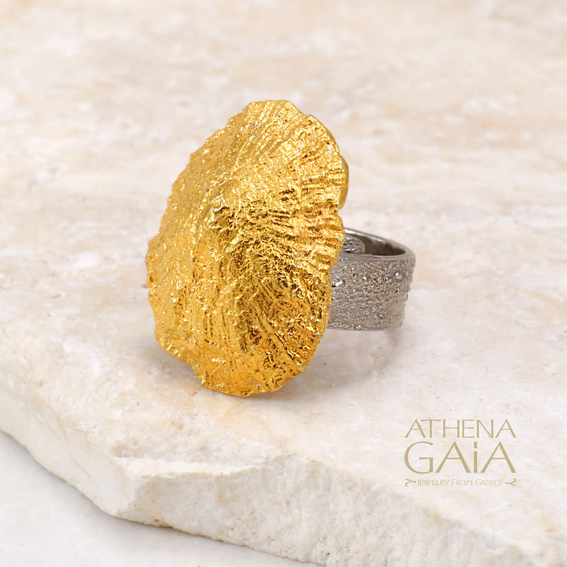A gold-colored seashell ring displayed on a stone surface with the brand name Athena Gaia visible.