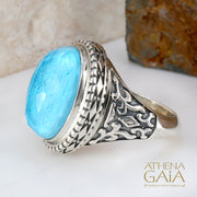 A silver-colored metal ring with a large blue stone set in the center, featuring intricate embossed patterns around the band.
