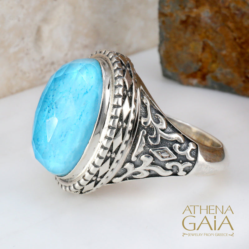 A silver-colored metal ring with a large blue stone set in the center, featuring intricate embossed patterns around the band.