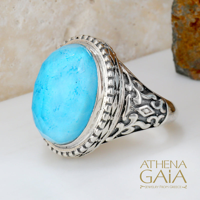 A silver ring with a large turquoise doublet, featuring intricate border details, displayed on a white surface with a textured background.