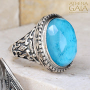 A silver ring with a large turquoise doublet, featuring intricate border details, displayed on a white surface with a textured background.