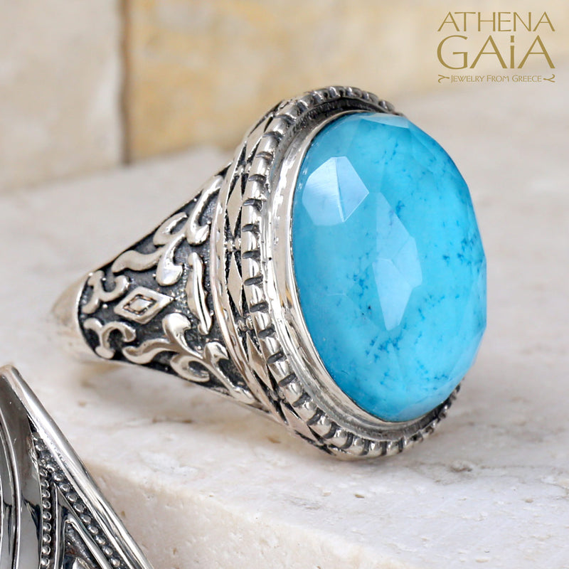 A silver ring with a large turquoise doublet, featuring intricate border details, displayed on a white surface with a textured background.