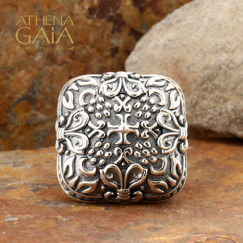 A silver-colored unisex ring with a floral pattern design, displayed on a textured background.