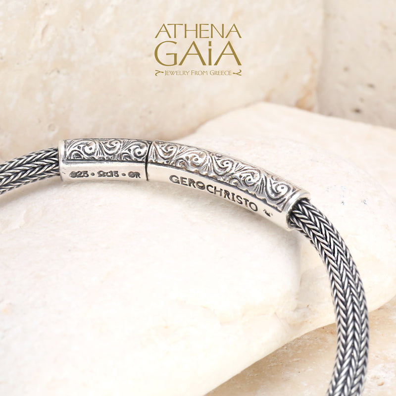 Handwoven silver bracelet with ornate scroll clasp detail, displayed on smooth beige stone.